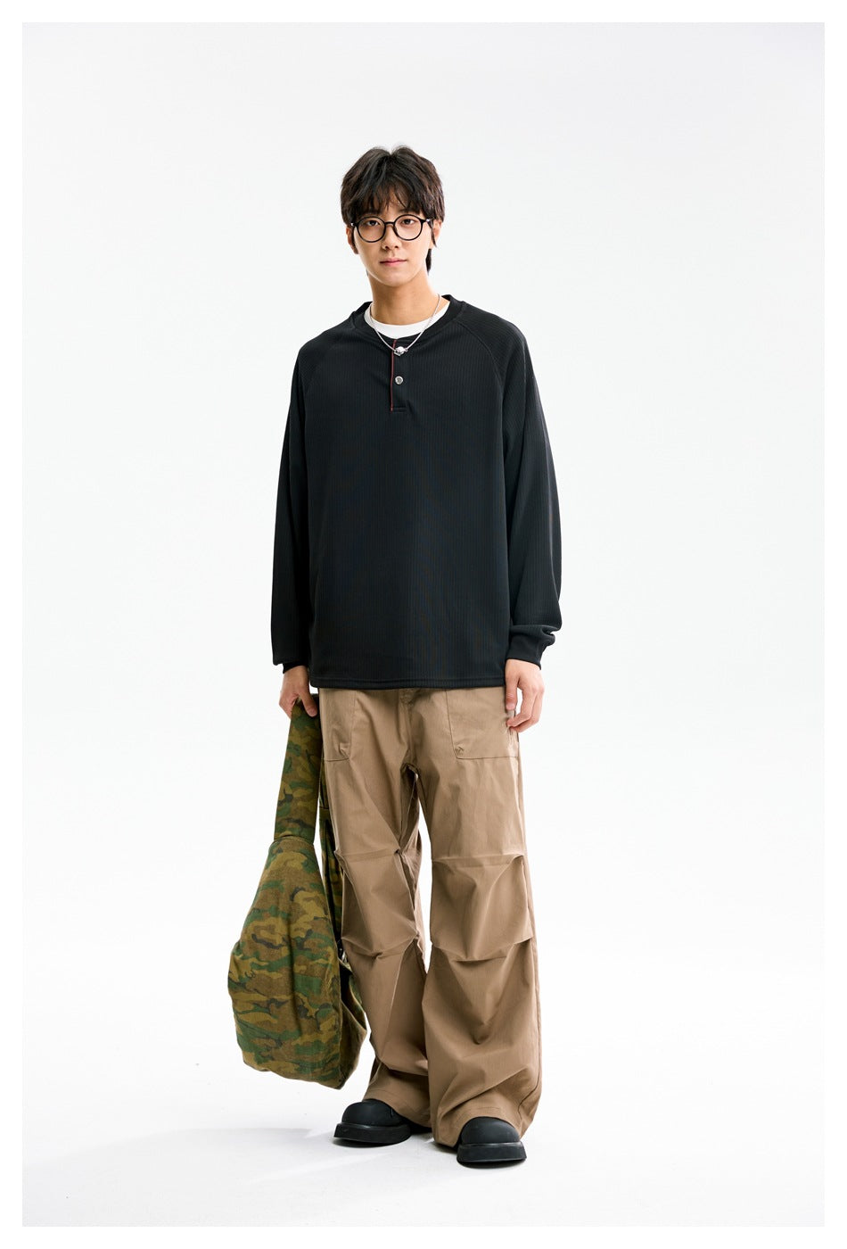 Front view of a model wearing a black long-sleeve henley with a short button placket and loose tan pants.