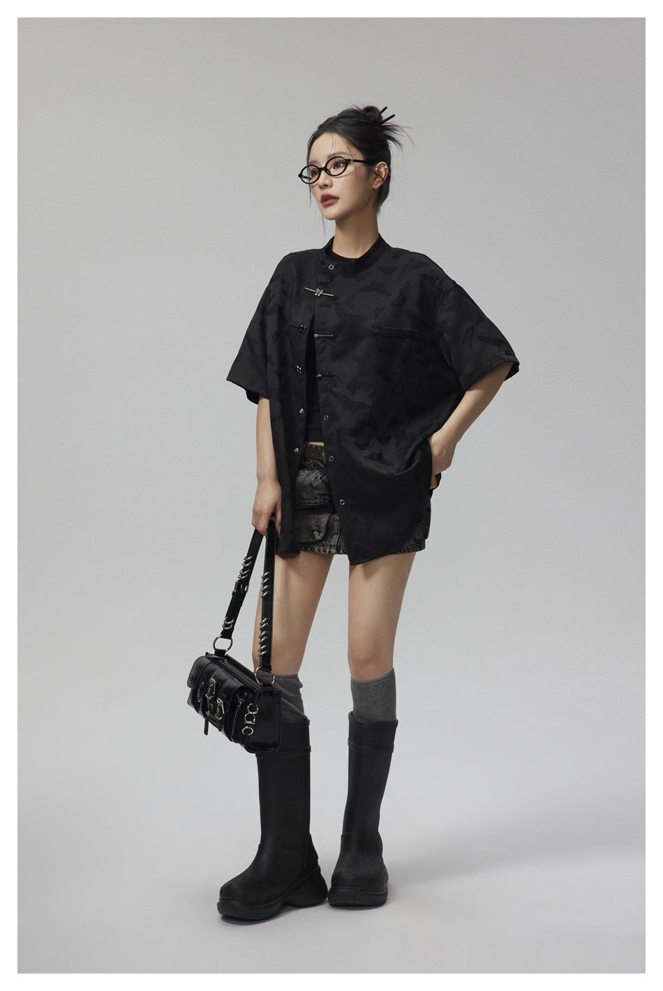 Model wearing the black butterfly-pattern short-sleeve shirt open over a dark top with denim skirt, boots and black shoulder