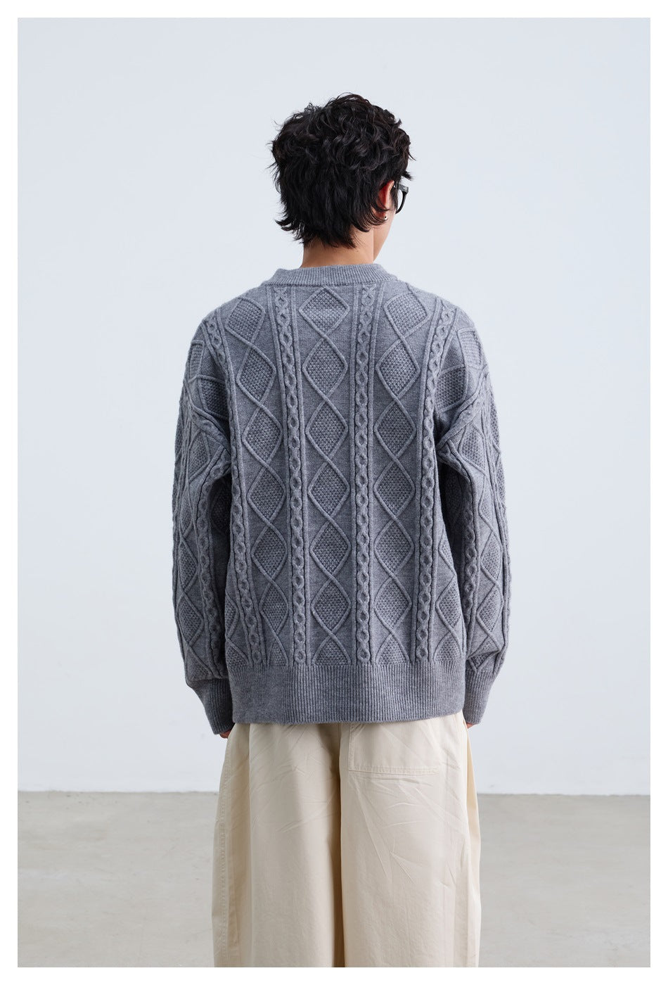 Back view of a model wearing a gray cable-knit sweater with vertical cable and diamond patterns and a ribbed hem.