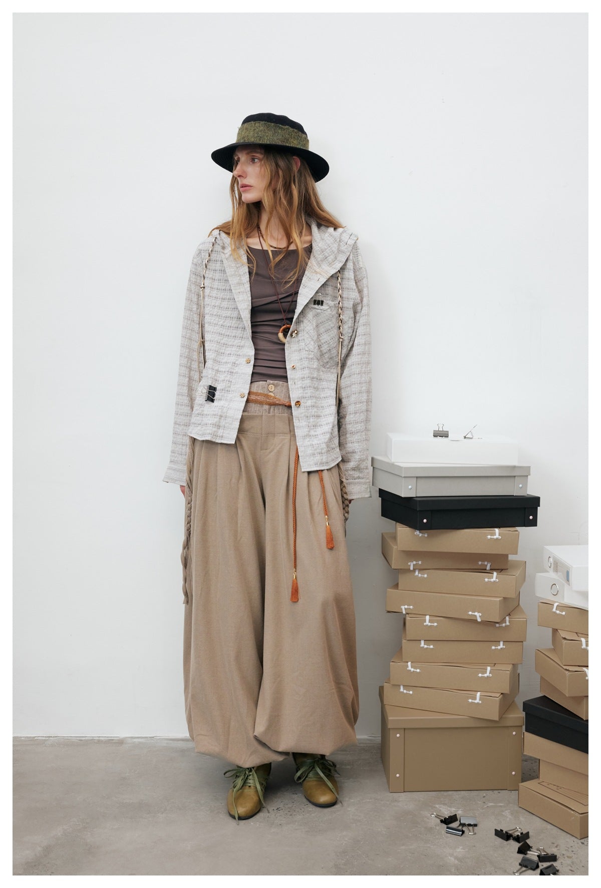 Full-length model wearing a light gray plaid hooded jacket with wide beige trousers and a dark hat.
