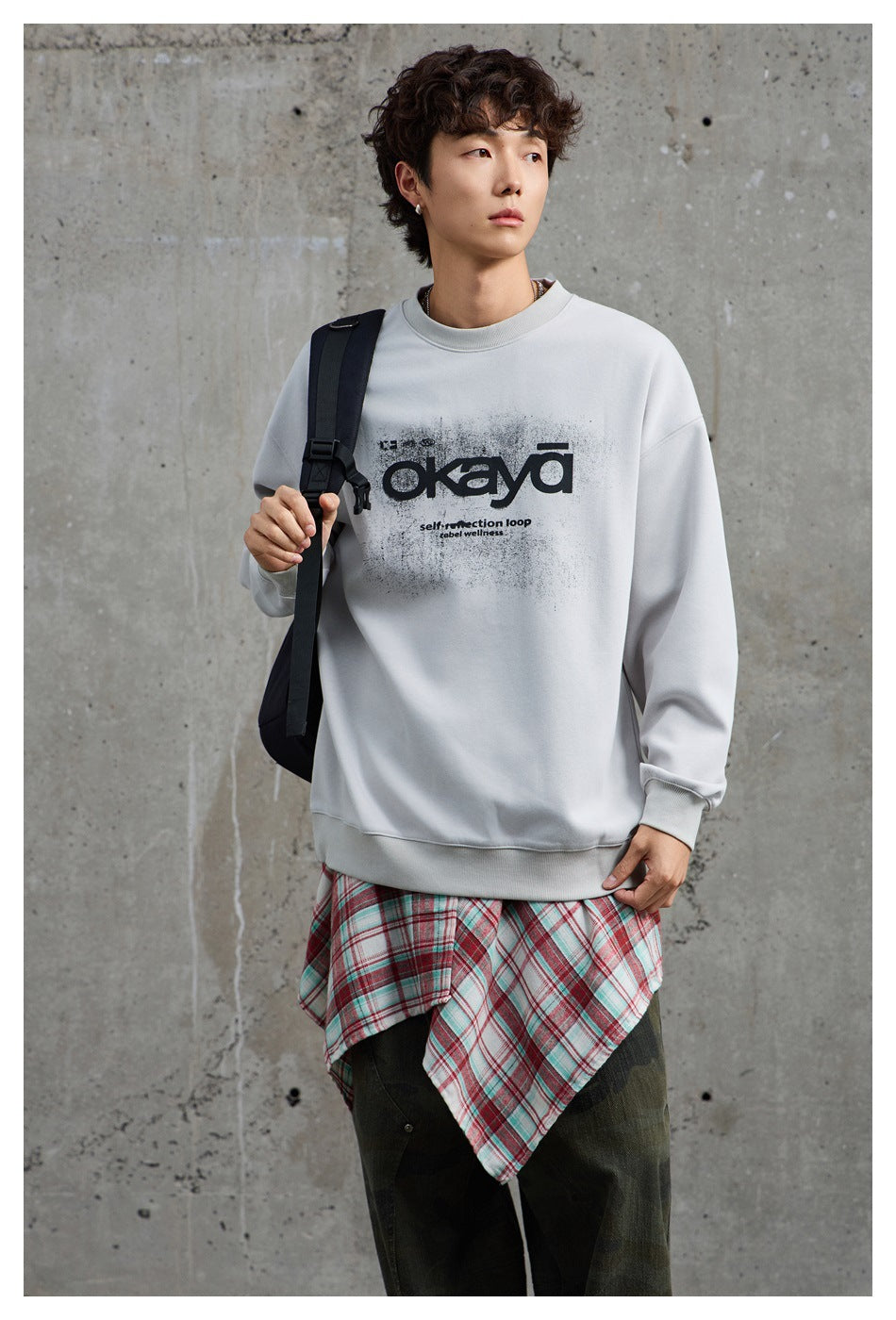 Model wearing a light gray graphic crewneck sweatshirt with a backpack and plaid shirt tied at the waist.