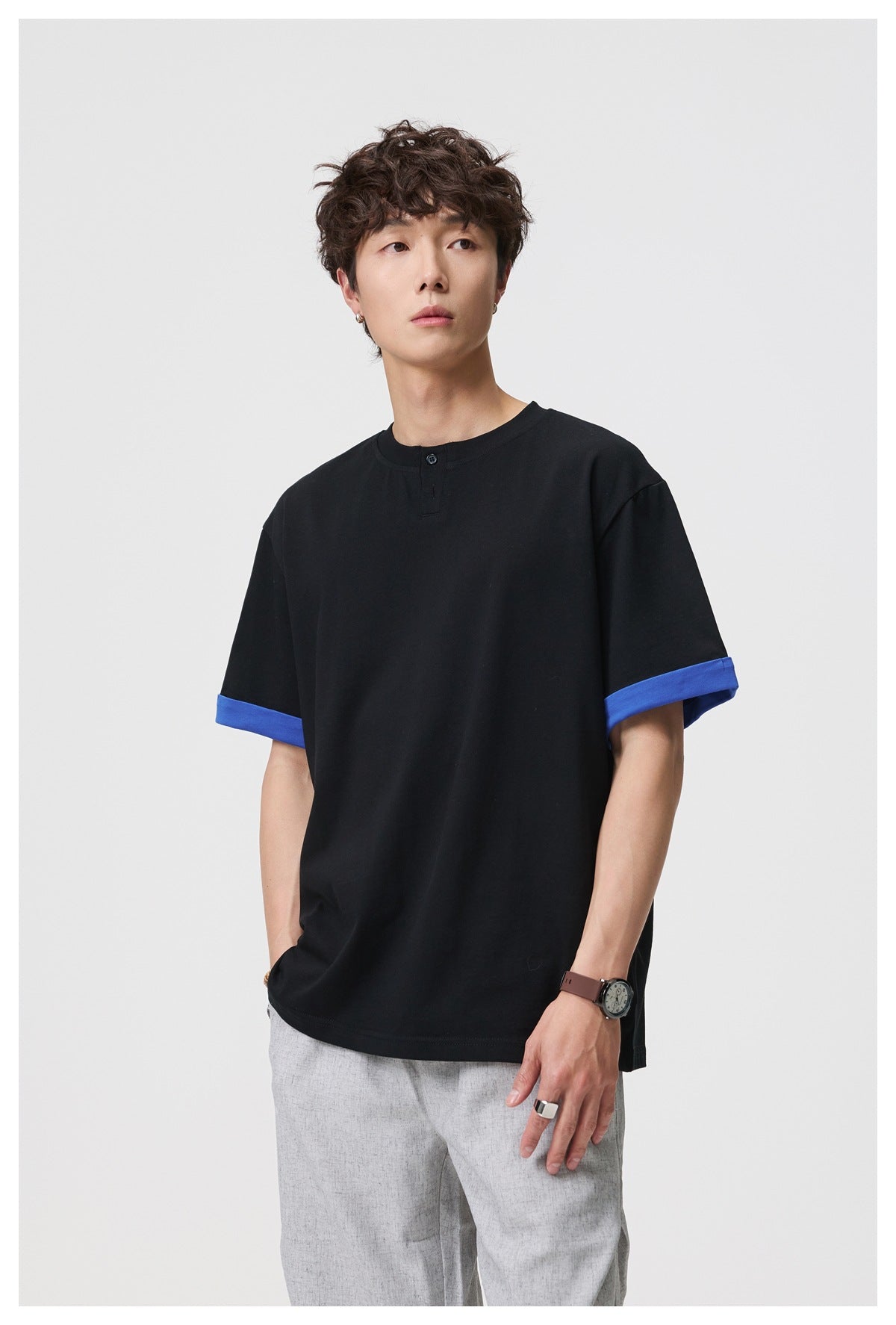Front view of a model wearing a black henley T-shirt with blue turned-up sleeve cuffs and light gray trousers.