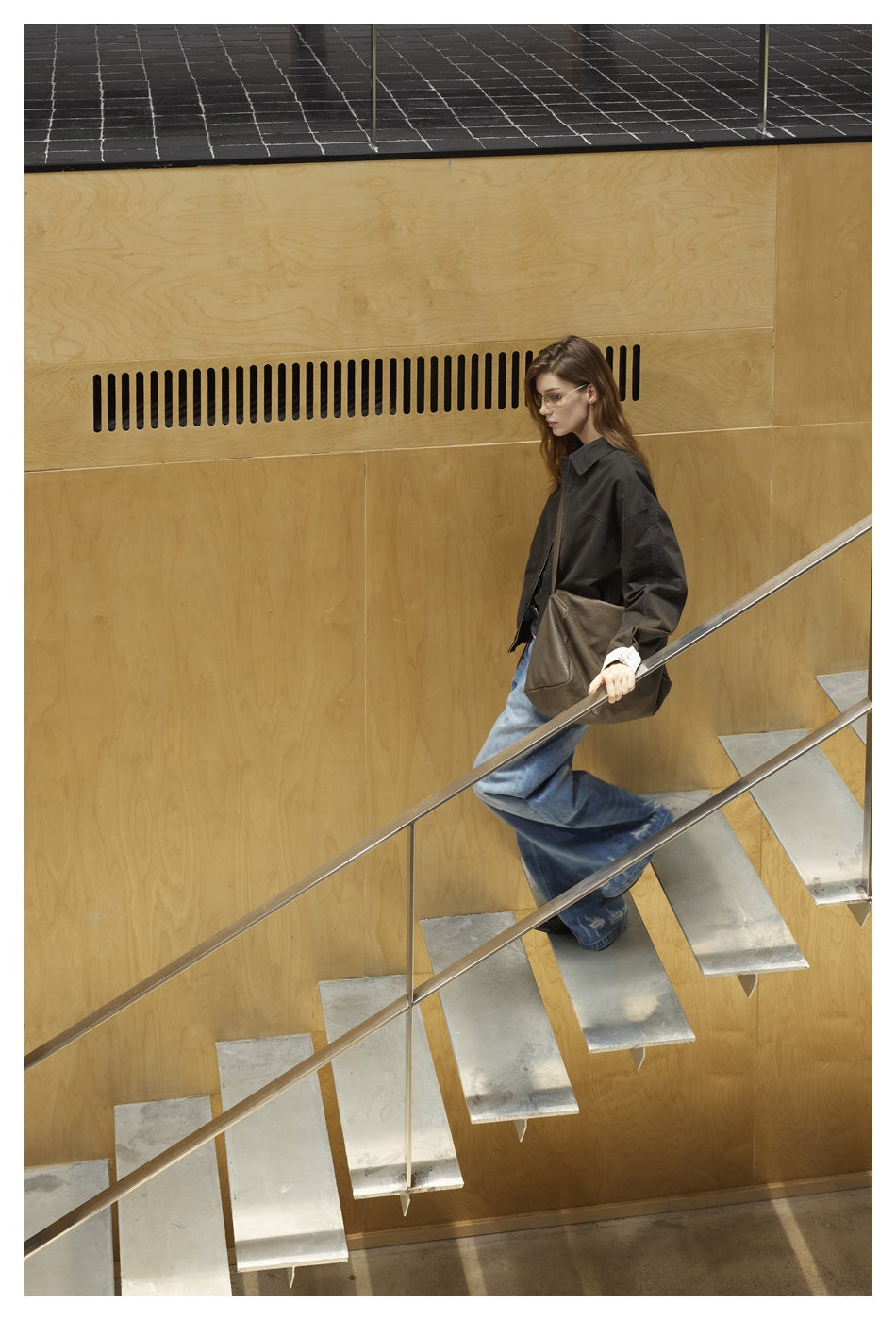 Model wearing a charcoal jacket and wide-leg blue jeans while walking on a staircase, with a brown shoulder bag.