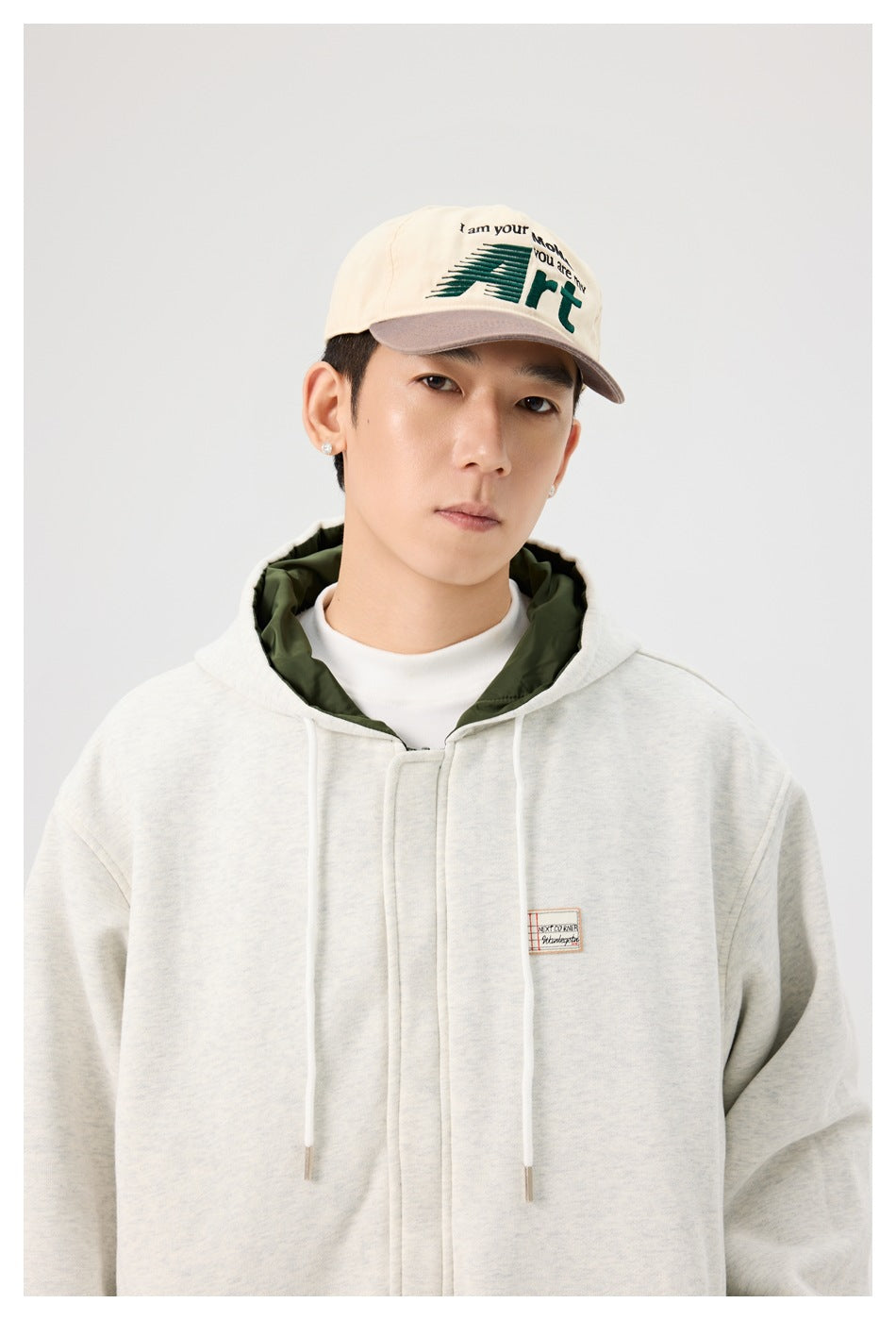 Man in a gray hooded zip jacket with white drawstrings, green lining at the hood, wearing a cap and white top