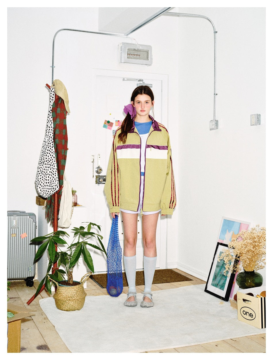 Model wearing an oversized olive green track jacket with plum and cream chest band over a blue top and shorts
