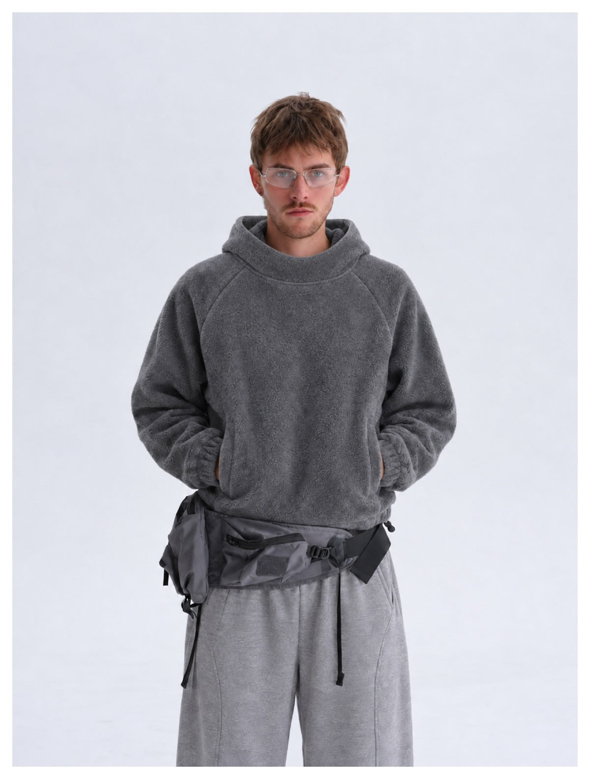 Front view of a model in a dark gray textured hooded sweatshirt and light gray pants, wearing a dark utility bag at the