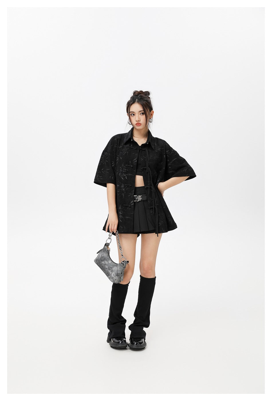 Model posing in a black short-sleeve patterned tie-front shirt, crop top and pleated skirt, with a silver shoulder bag.