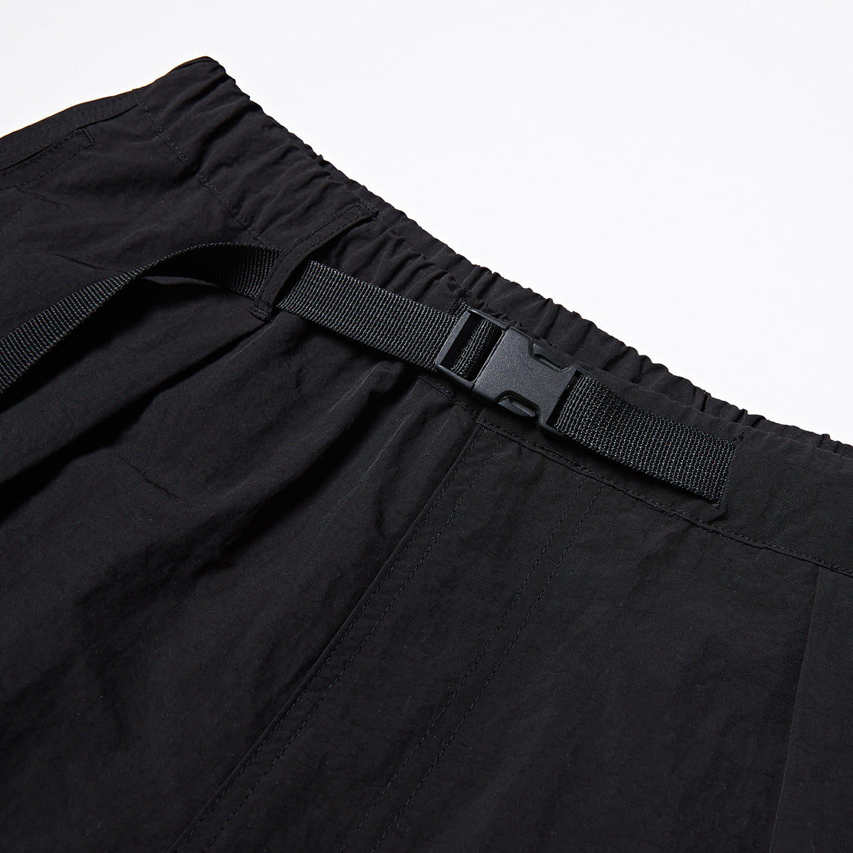 Close-up of black shorts showing an elastic waistband, webbing belt, plastic buckle and stitched panels.
