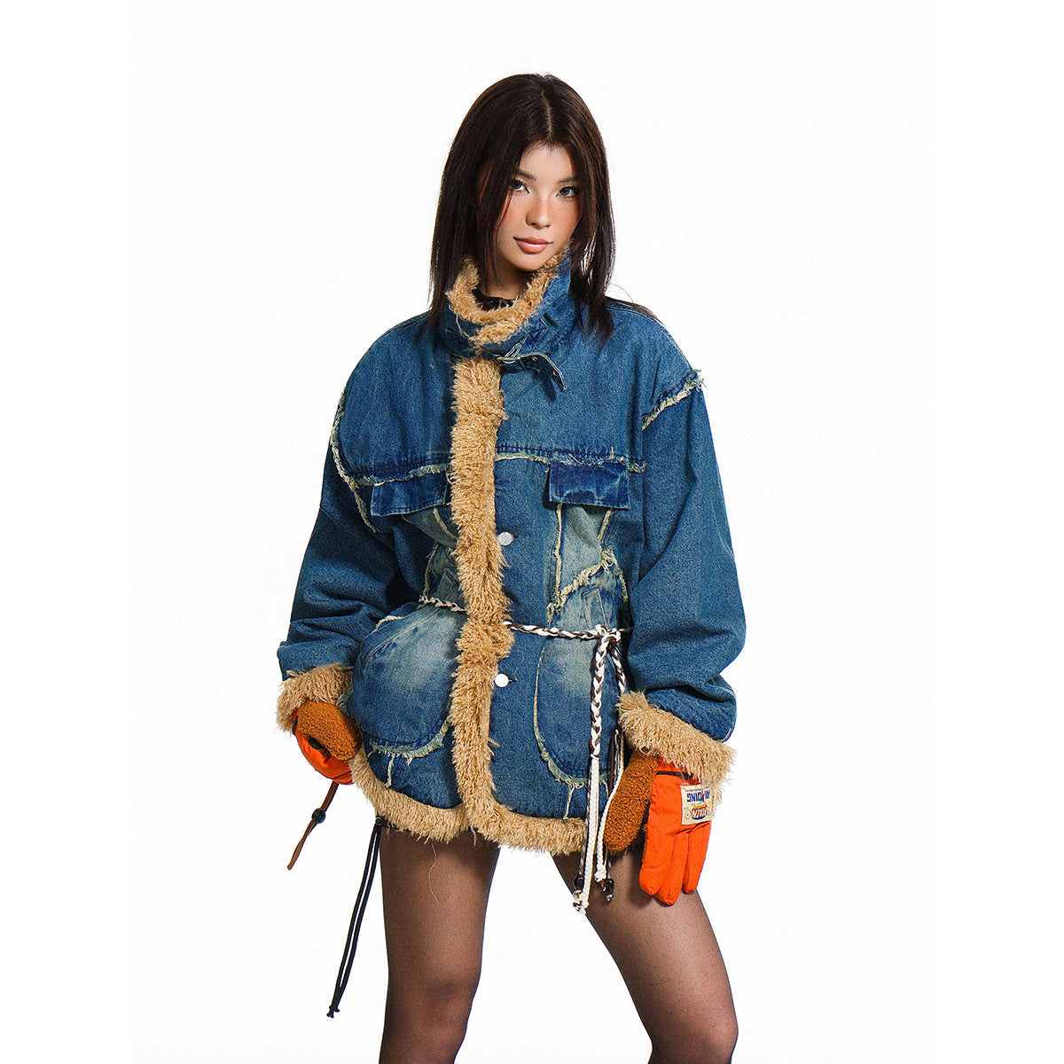 Model wearing an oversized blue patchwork denim jacket with tan shaggy trim, a braided waist cord and orange gloves.