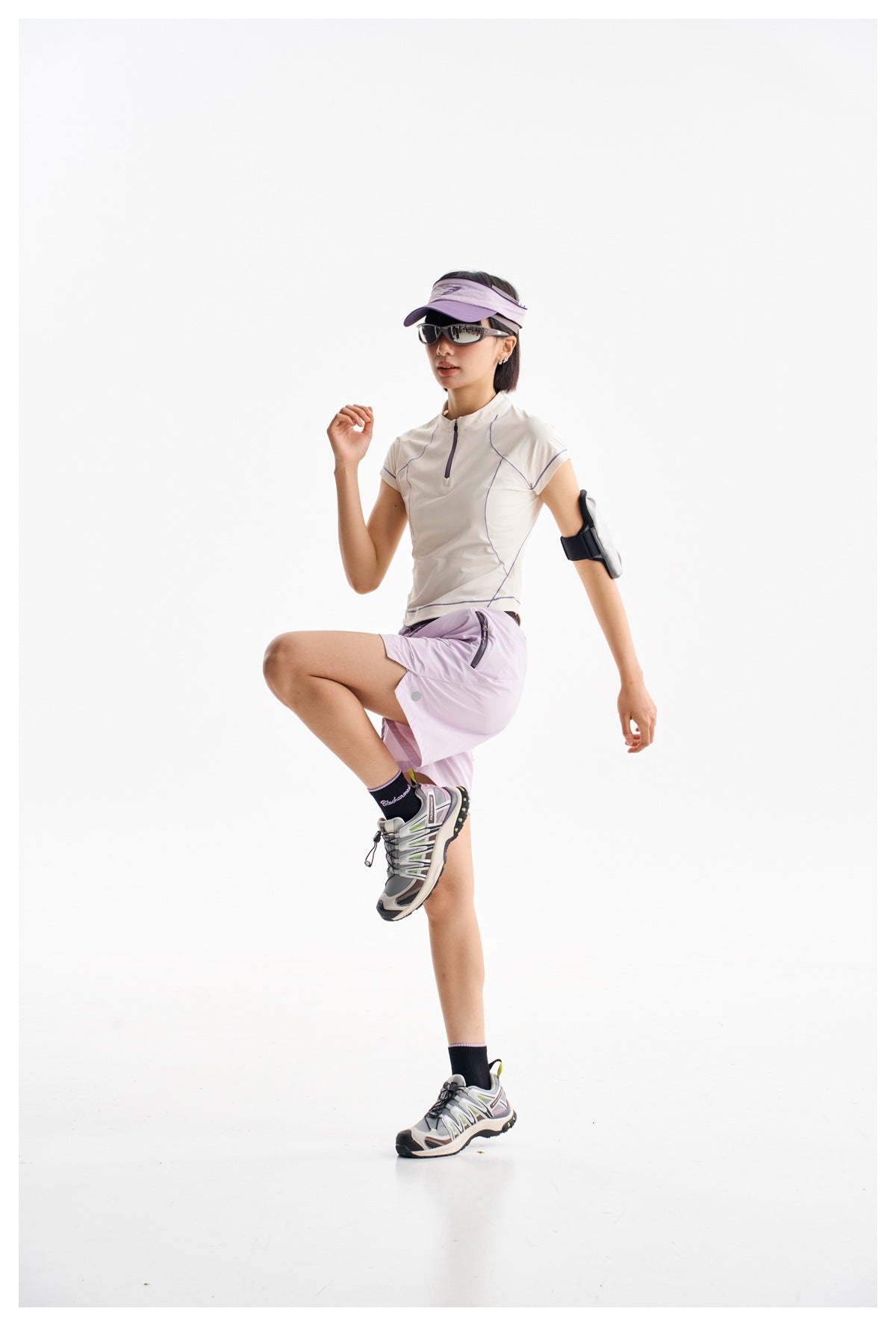 Model posing in a white quarter-zip top with purple piping and lavender shorts, visor, sunglasses, and running shoes.