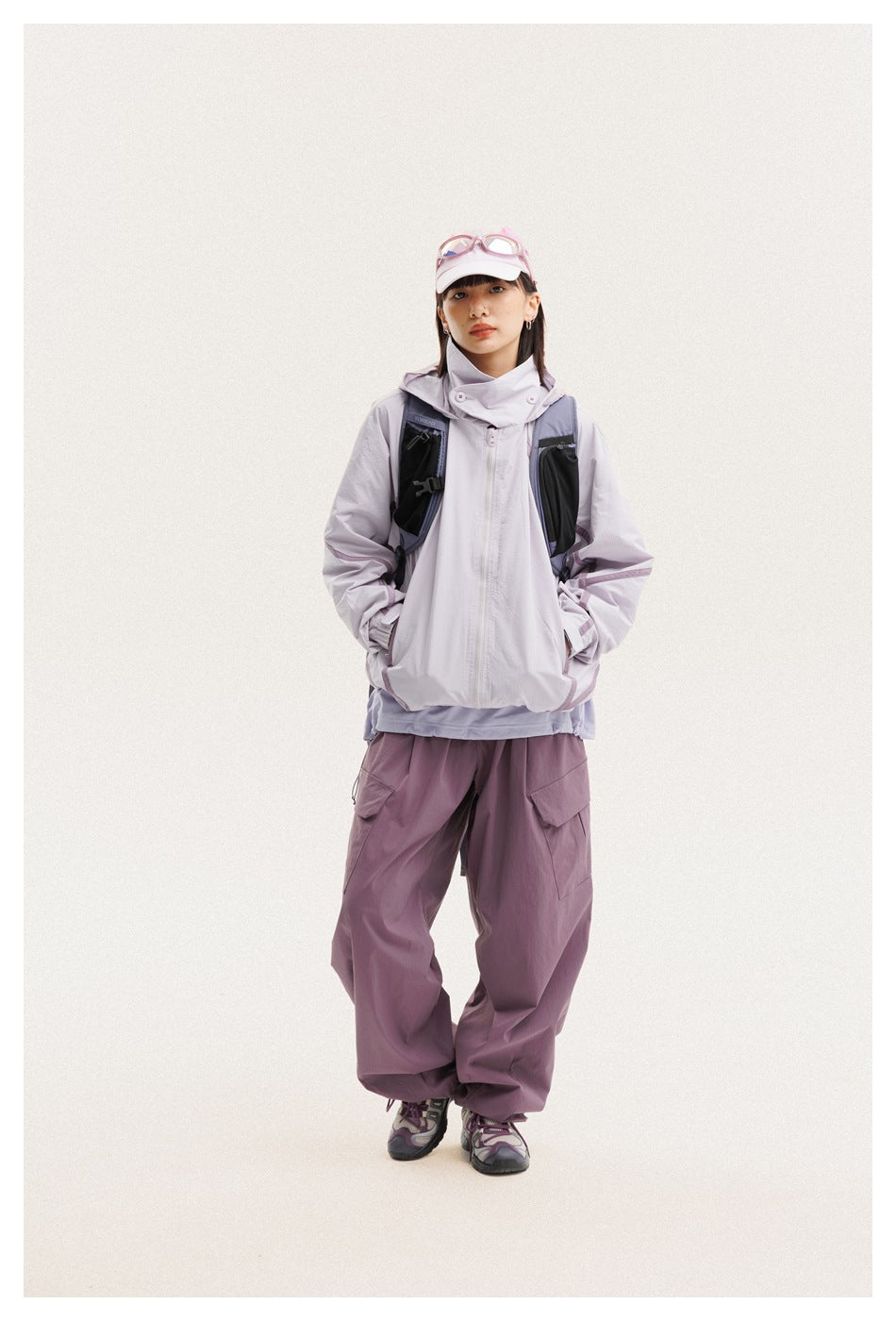 Model wearing a pale lavender hooded jacket, dark vest, and loose plum cargo pants with a pink cap.