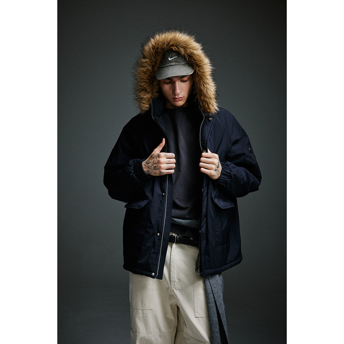 Model wearing a dark navy hooded parka with brown fur trim, layered over a dark top and beige trousers.
