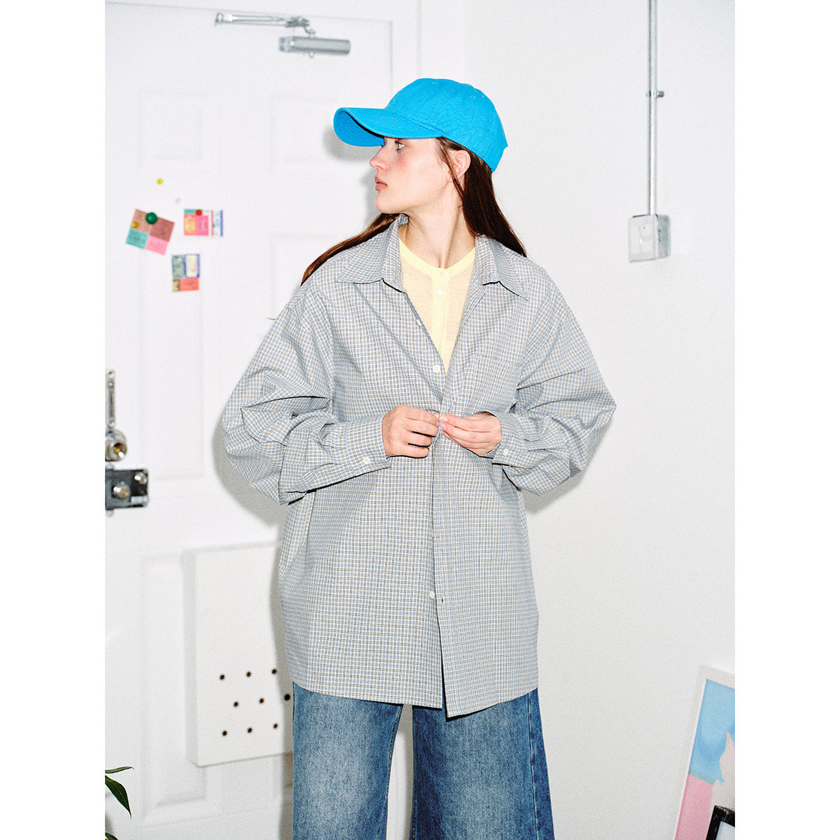 Front view of a model wearing an oversized pale blue check button-up shirt, yellow top, jeans and a bright blue cap.