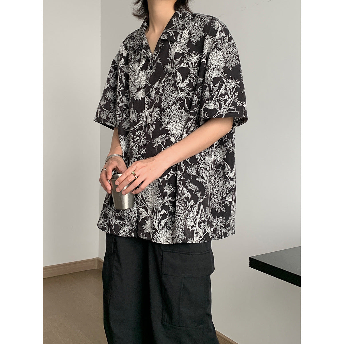 Model wearing an oversized black short-sleeve button-up shirt with a dense white botanical print and black cargo trousers.