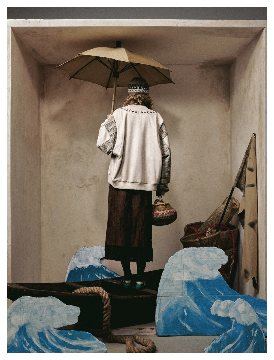 Back view of a model wearing an ivory jacket with patterned sleeves and a dark skirt, holding an umbrella and woven bag.