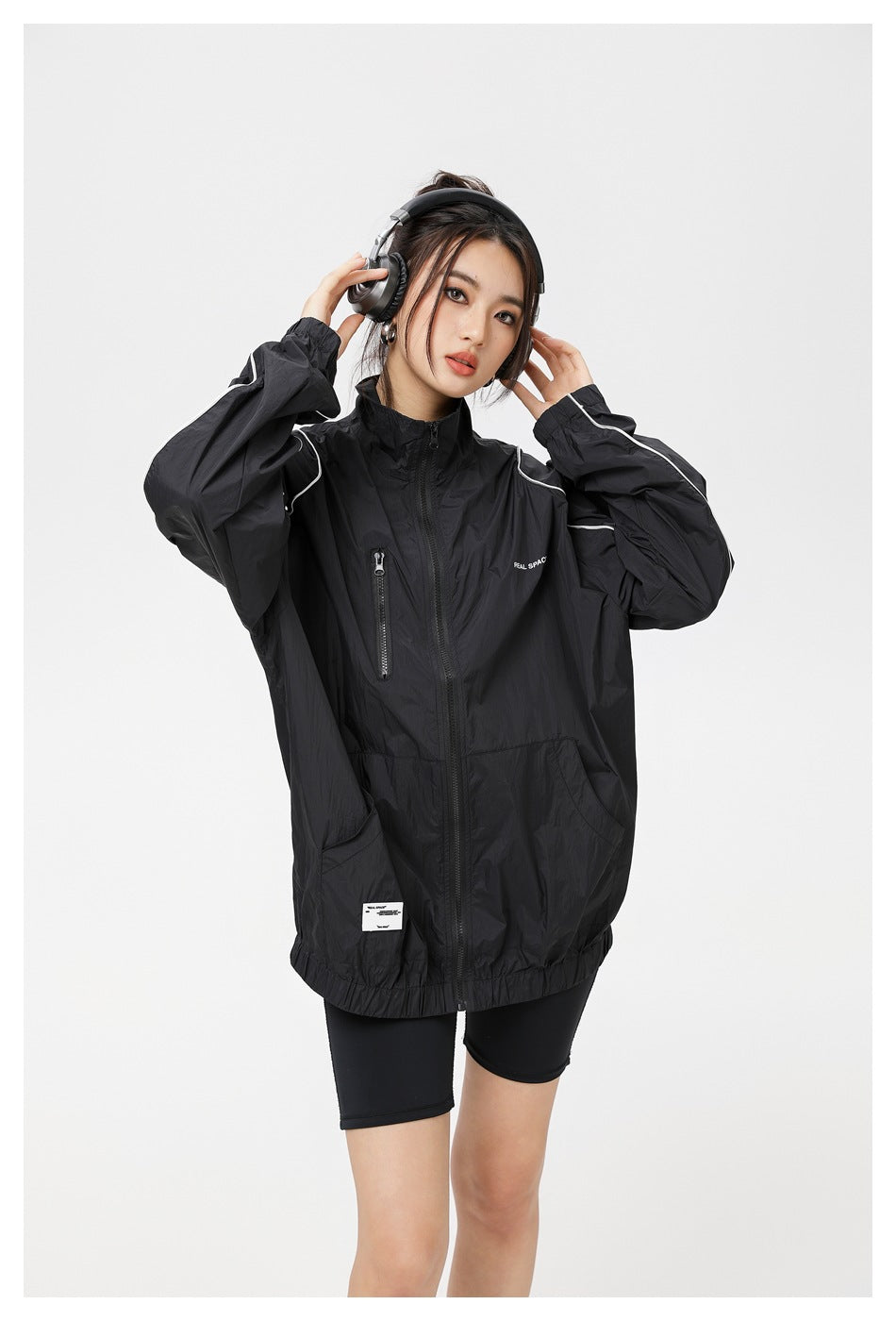 Model wearing an oversized black zip-up jacket with white piping, a high collar and front pockets.