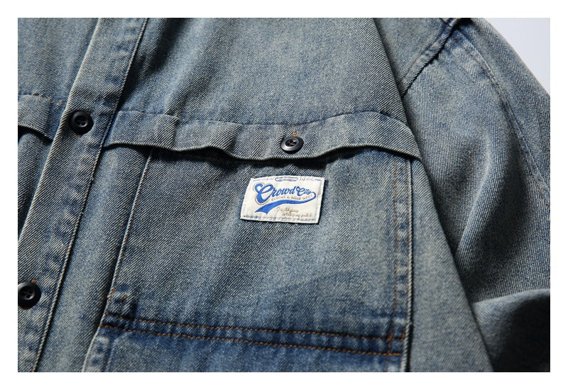 Close-up of a small sewn label on the gray denim shirt’s front pocket beneath a buttoned flap.