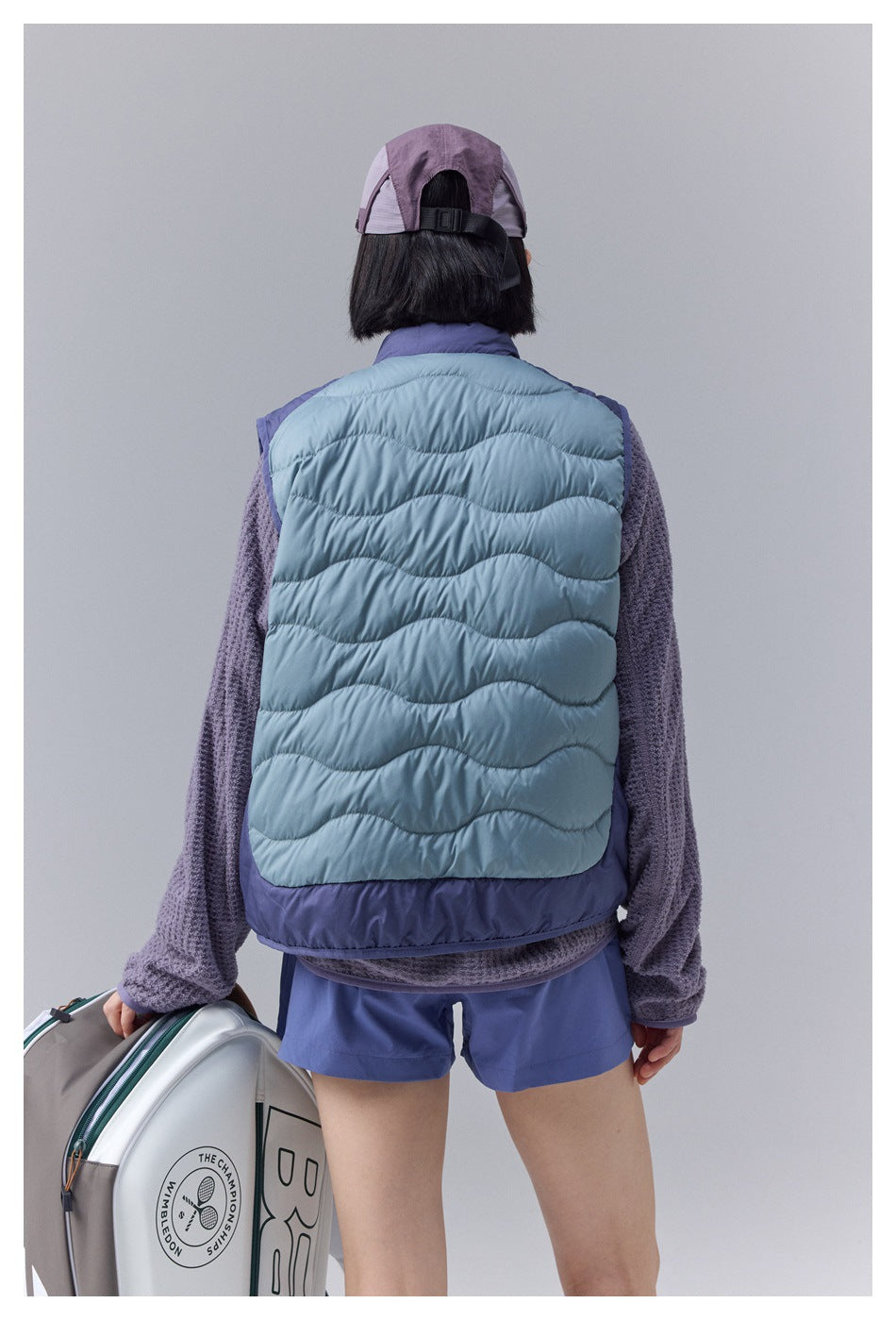 Rear view of a model wearing a pale blue quilted vest with wavy horizontal stitching and a dark purple hem.
