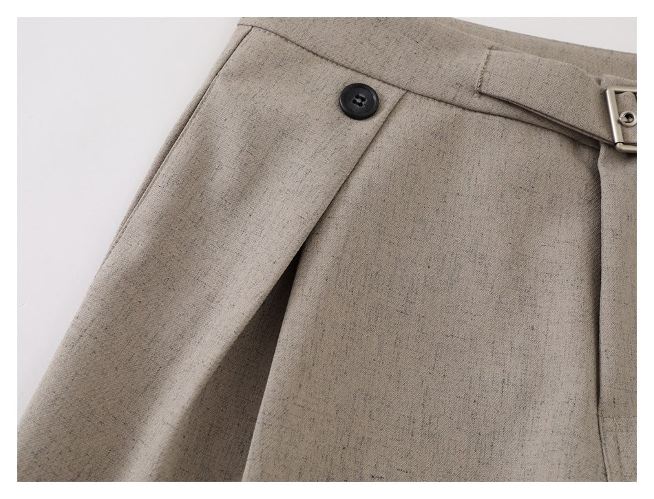 Close-up of taupe trousers showing a black waist button and overlapping front pleat.