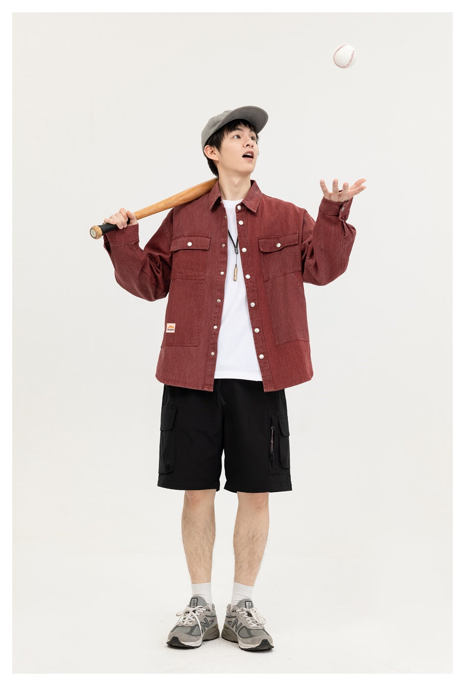 A man wears a burgundy button-front overshirt with chest and lower pockets, black cargo shorts and a gray cap.
