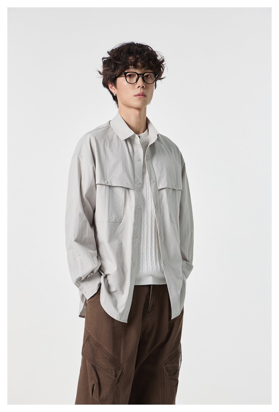Model wearing an open light gray flap-pocket shirt over a white cable-knit sweater with brown cargo trousers.