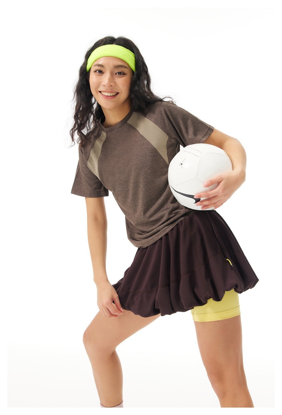 Model posing with a white ball in a brown paneled athletic top, dark flared skirt, yellow shorts and neon headband.
