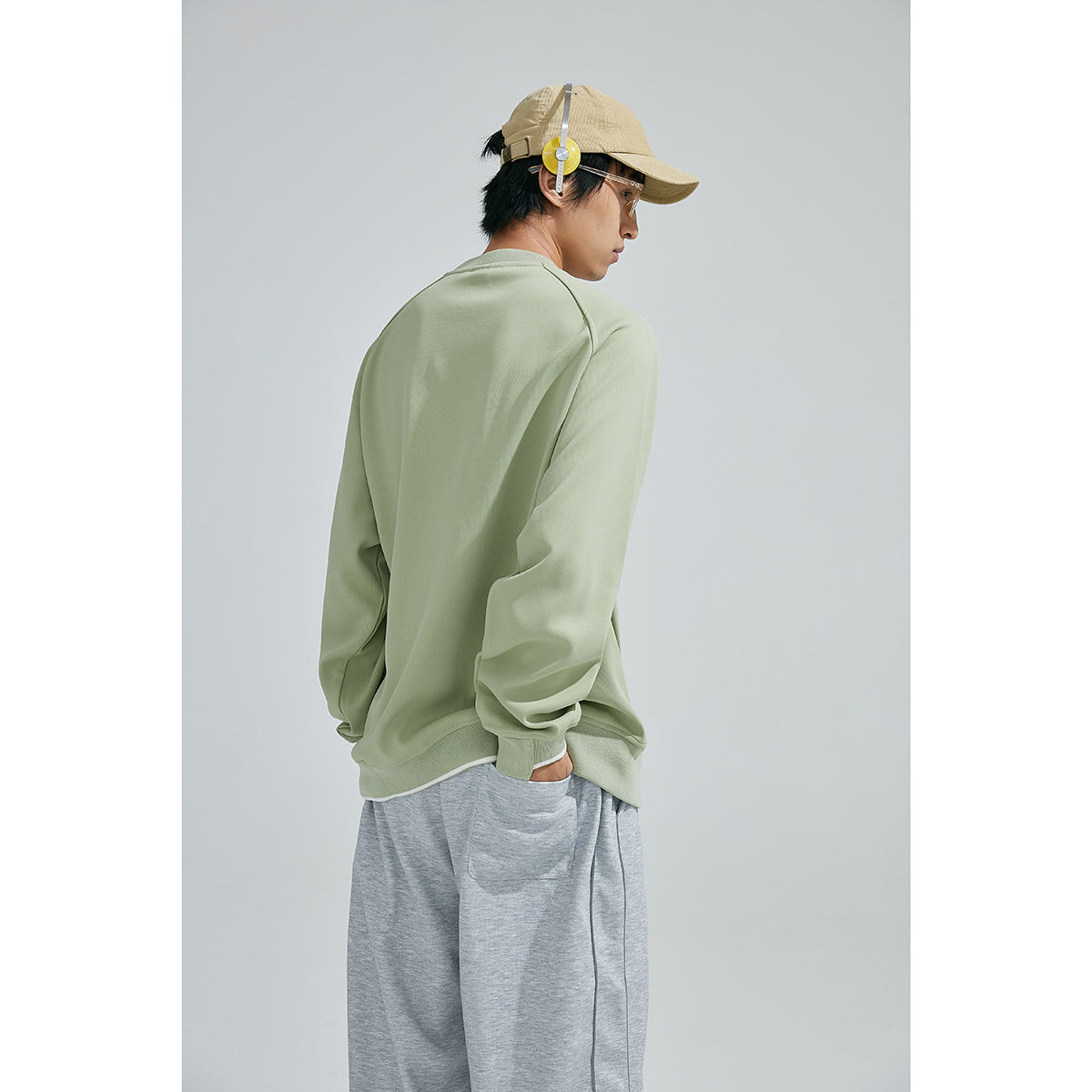 Three-quarter rear view of a model wearing a plain sage green sweatshirt and loose gray pants.