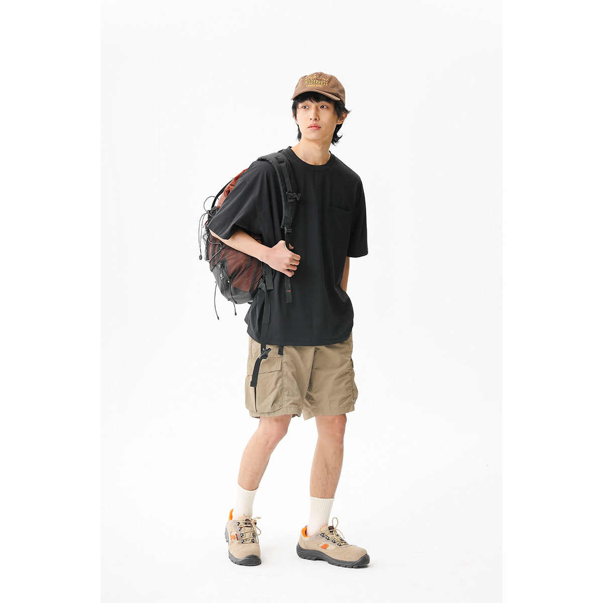 Man wearing a black pocket T-shirt and tan cargo shorts with a hiking backpack against a white background.