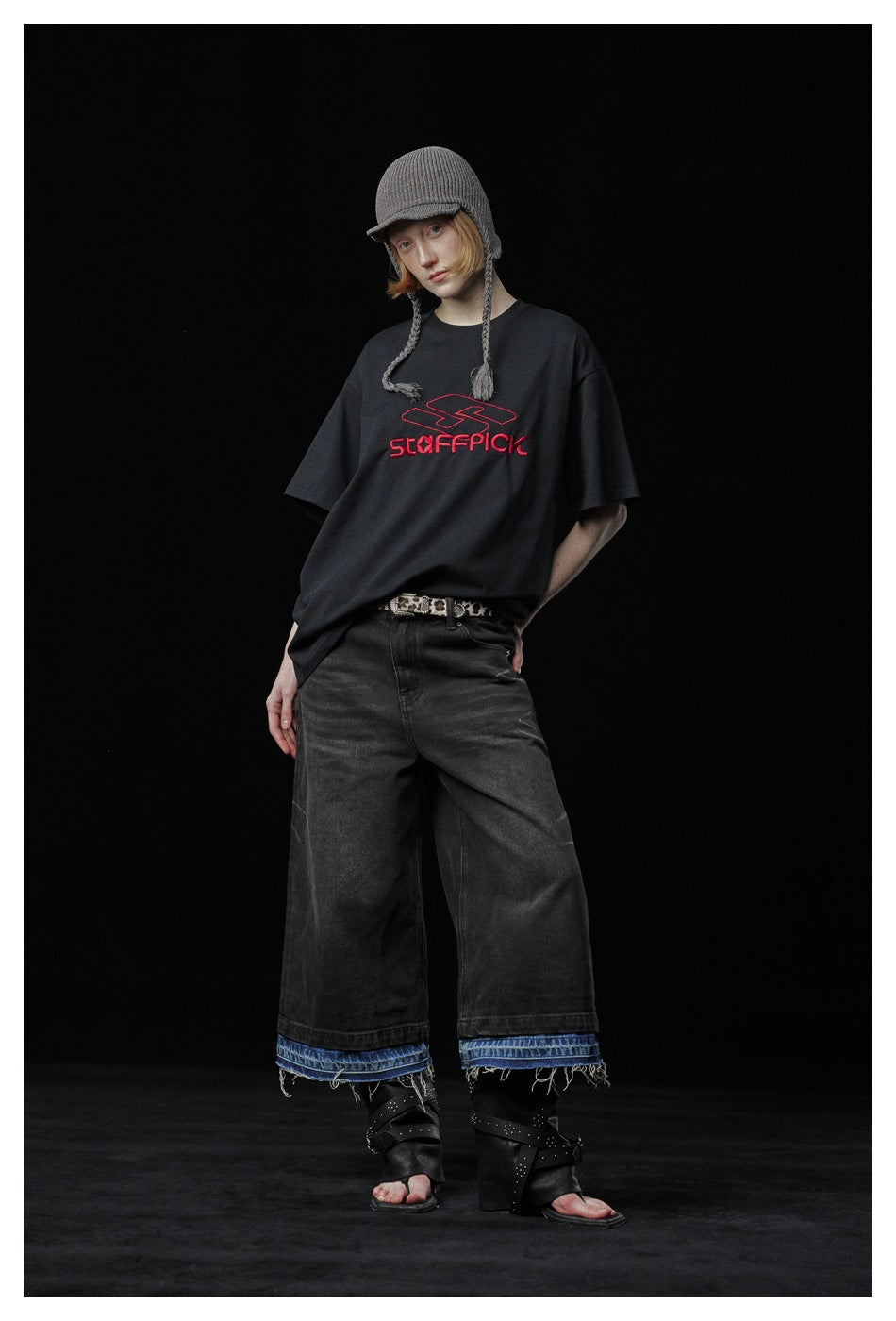 Full-length view of a model in a black T-shirt with red chest lettering, wide dark jeans, and a gray knit cap.