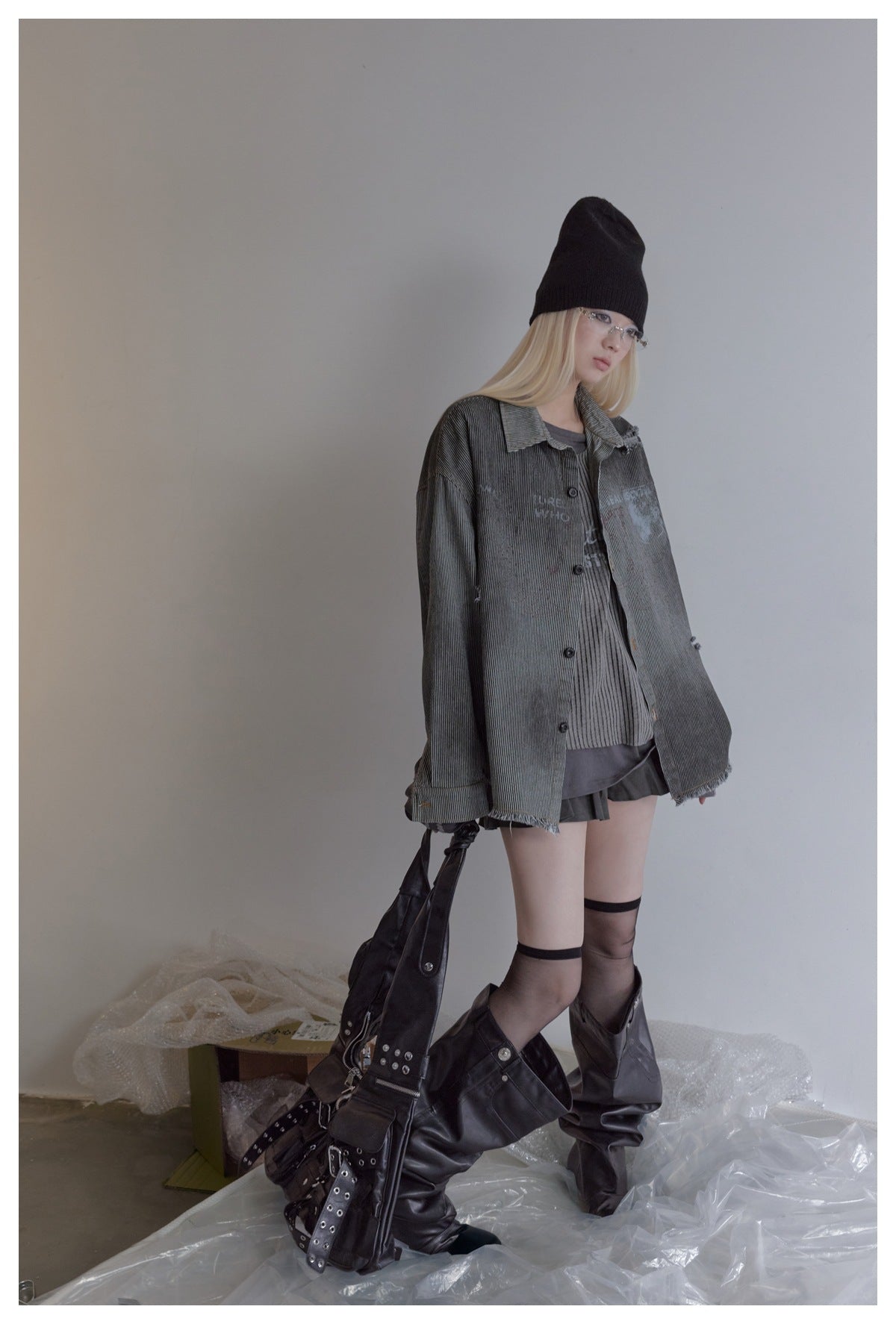 Model wearing an oversized charcoal pinstriped jacket with frayed hems, styled with a beanie and tall boots.