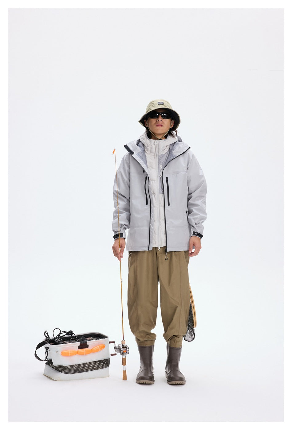 Full-length model wearing a light gray hooded jacket over a white layer with loose olive trousers and fishing gear.