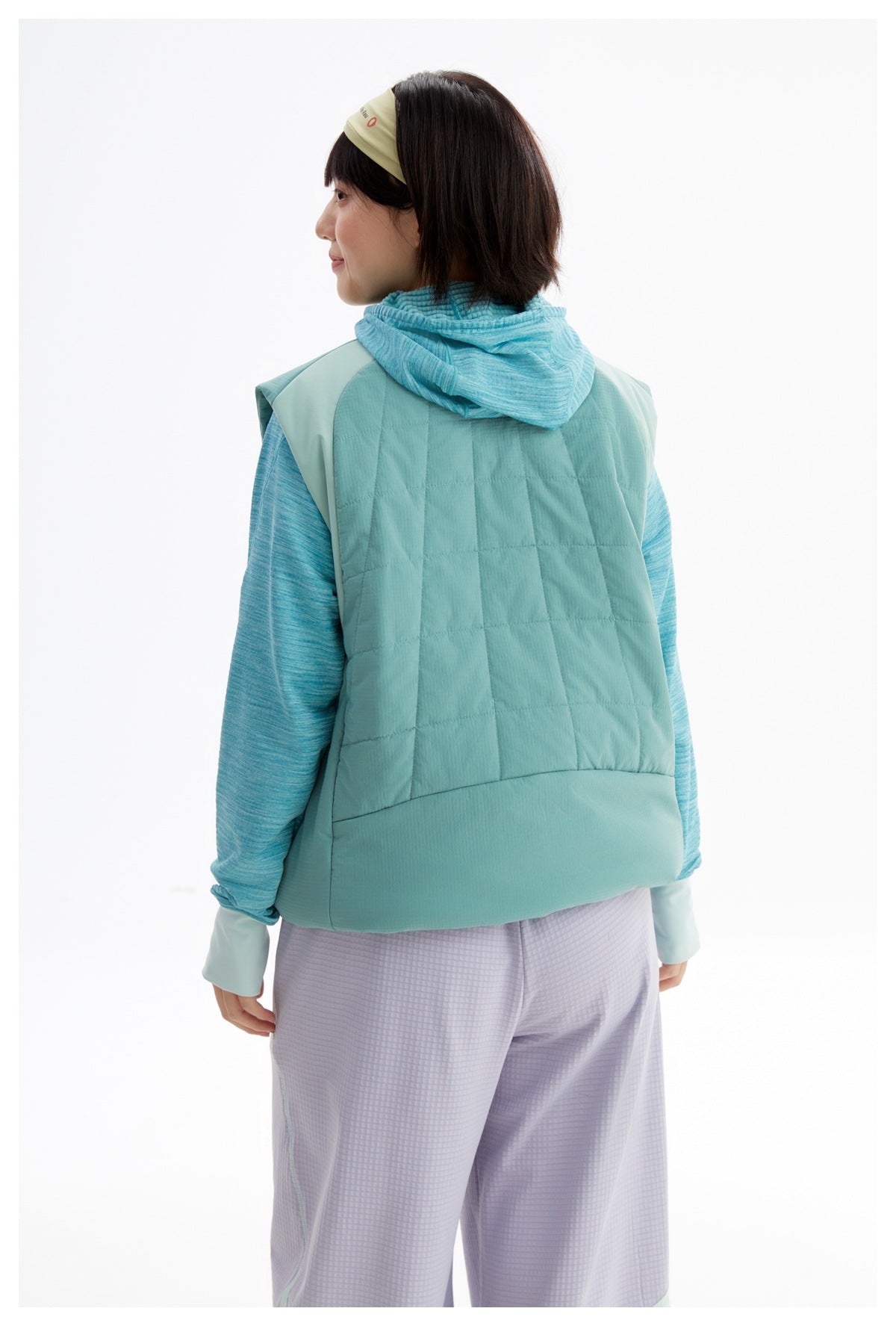 Back view of a model wearing a teal quilted vest with a hood and turquoise sleeves.