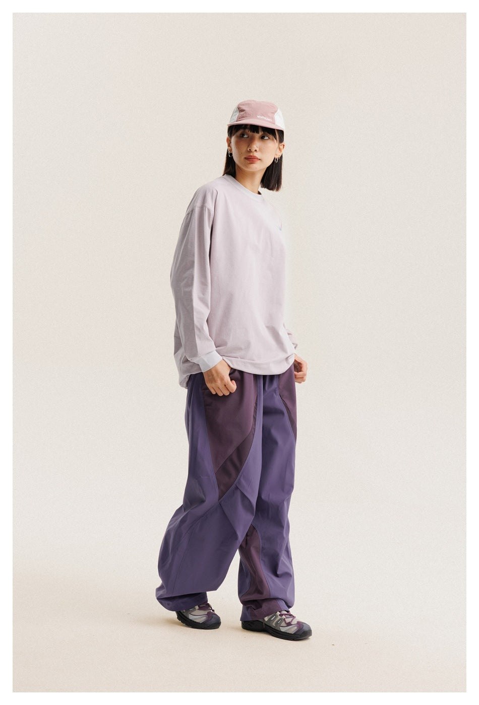 Model in a pale lilac long-sleeve shirt and loose purple paneled pants, shown at a three-quarter angle.