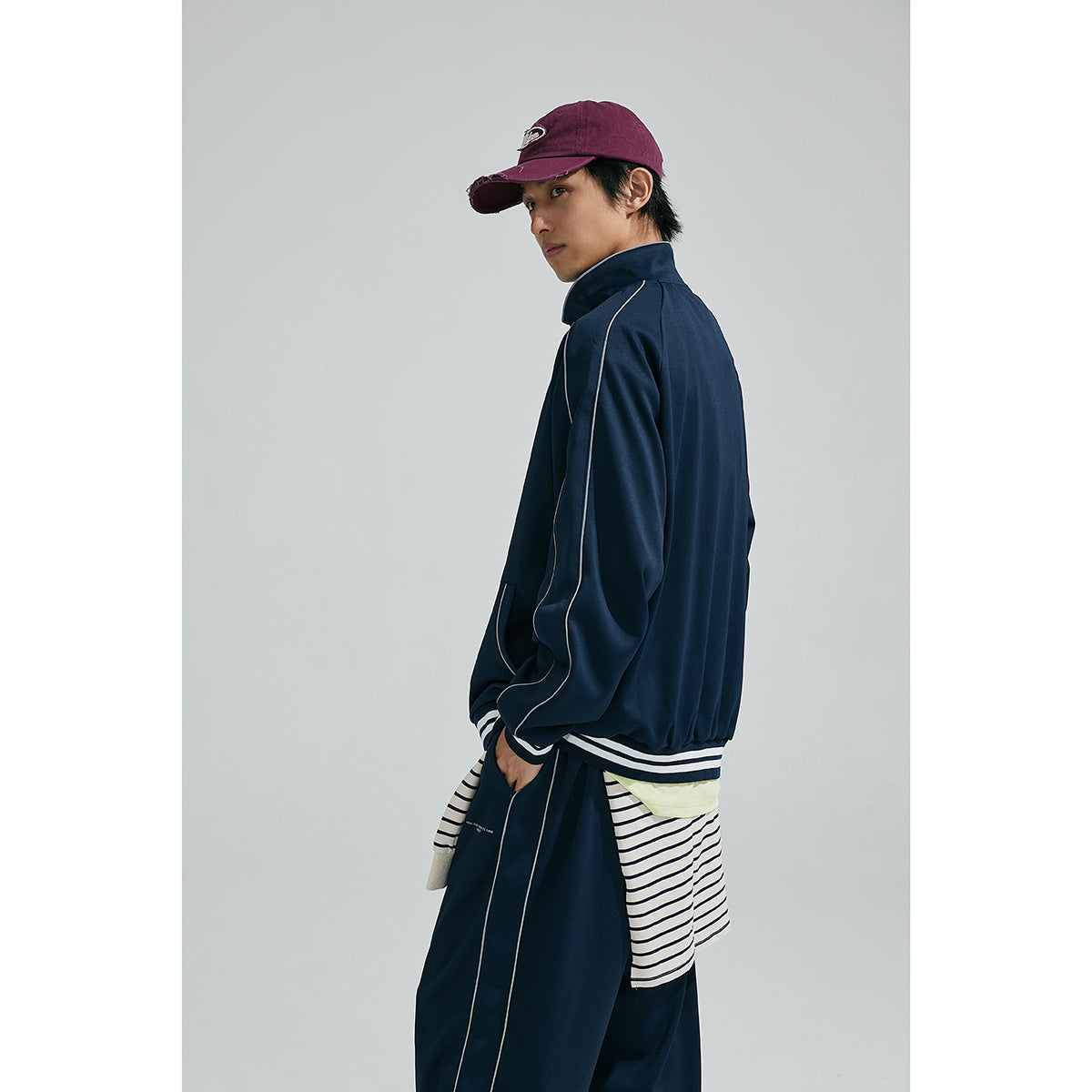 Model in a navy tracksuit shown from the side and back, with white piping and striped layers tied at the waist.