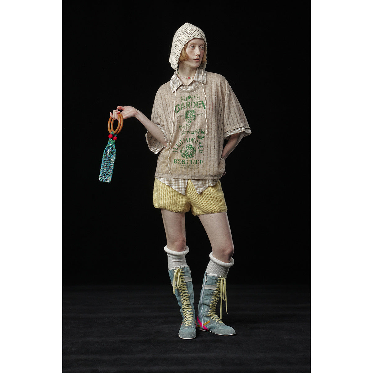 Model wearing a beige graphic tee over a checked shirt with yellow shorts, a cream crochet bonnet, and tall blue boots.