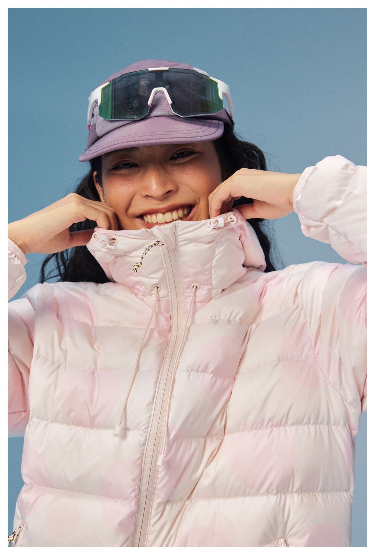 Close view of a smiling model in a pale pink hooded puffer jacket, adjusting its high collar.