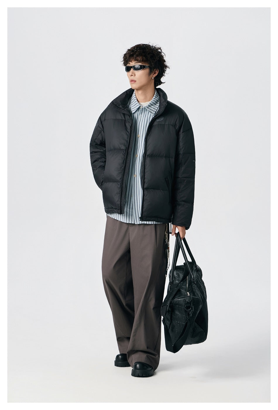 Model wearing a black quilted puffer jacket with a raised collar, striped shirt and wide dark trousers.