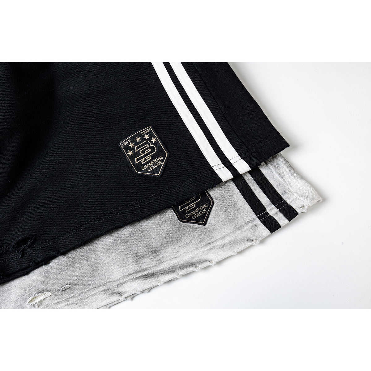 Close-up of black and gray athletic shorts hems with twin side stripes, embroidered shield patches, and distressed edges.