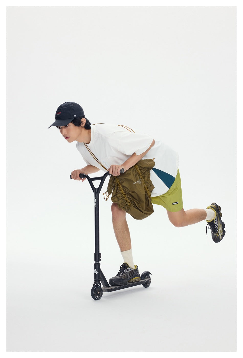 Model riding a scooter in green shorts with a white tee, black cap and an olive bag partly covering the shorts