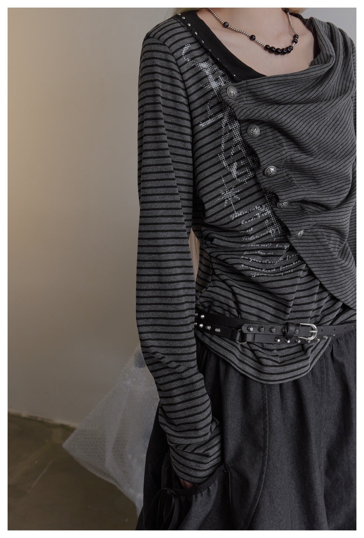 Close view of the striped top's draped front, decorative buttons, printed text, and studded belt on a model.