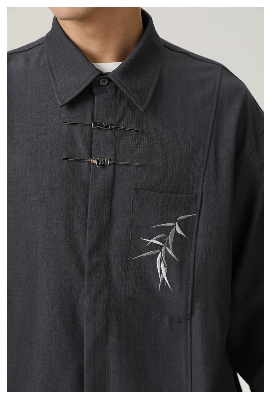 Close-up of a person wearing a charcoal shirt with a pointed collar, twin metal chest fasteners, and bamboo embroidery on the
