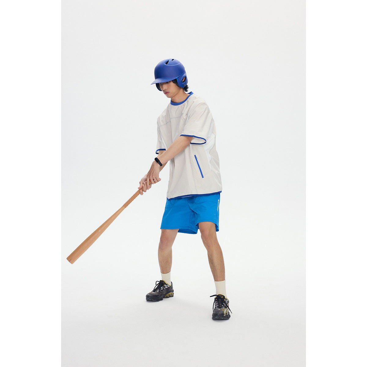 Model wearing an ivory short-sleeve shirt with blue trim and bright blue shorts, holding a bat and wearing a blue helmet.