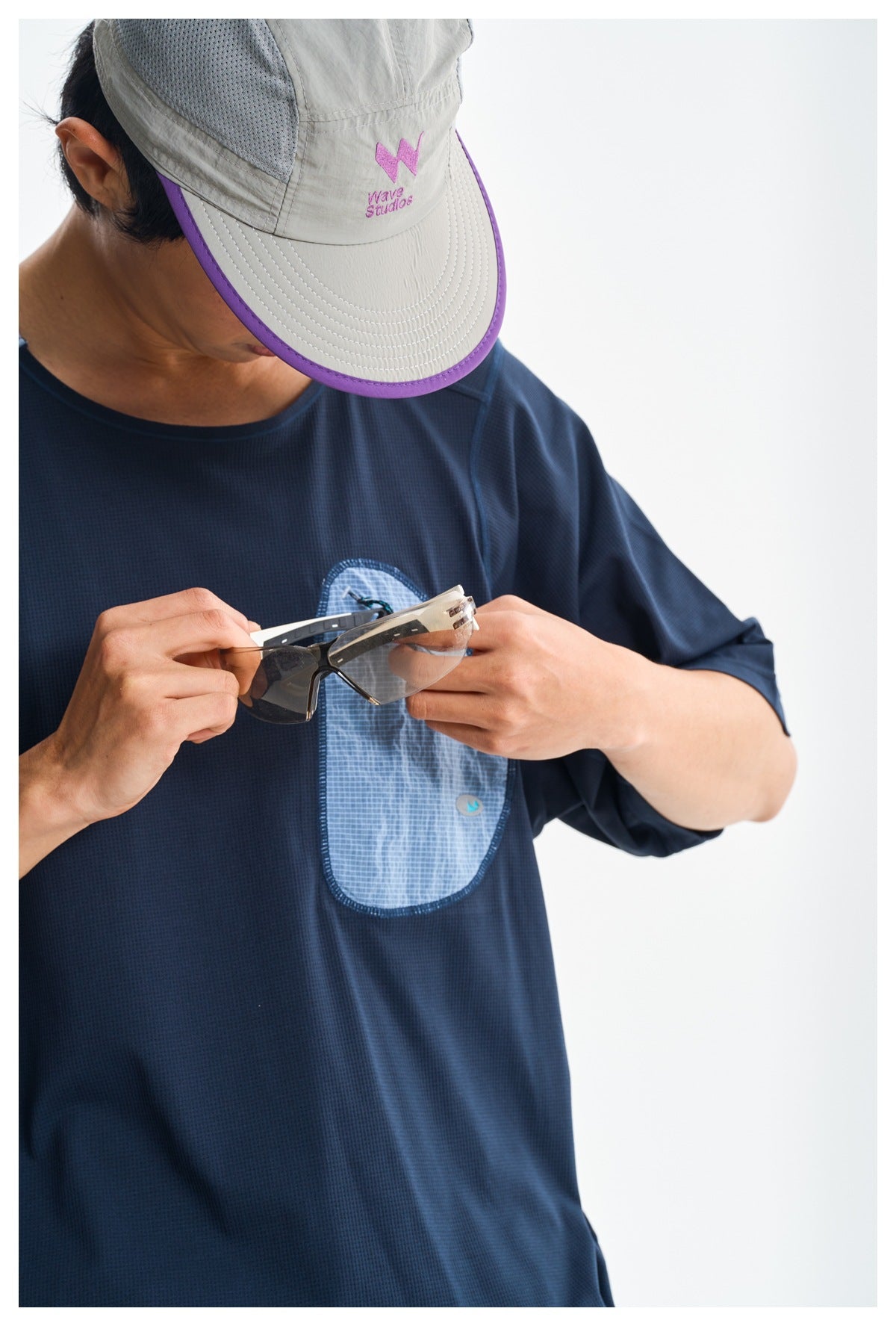 Close view of a navy shirt's light blue chest pocket as the model holds sunglasses; a gray cap is also visible.