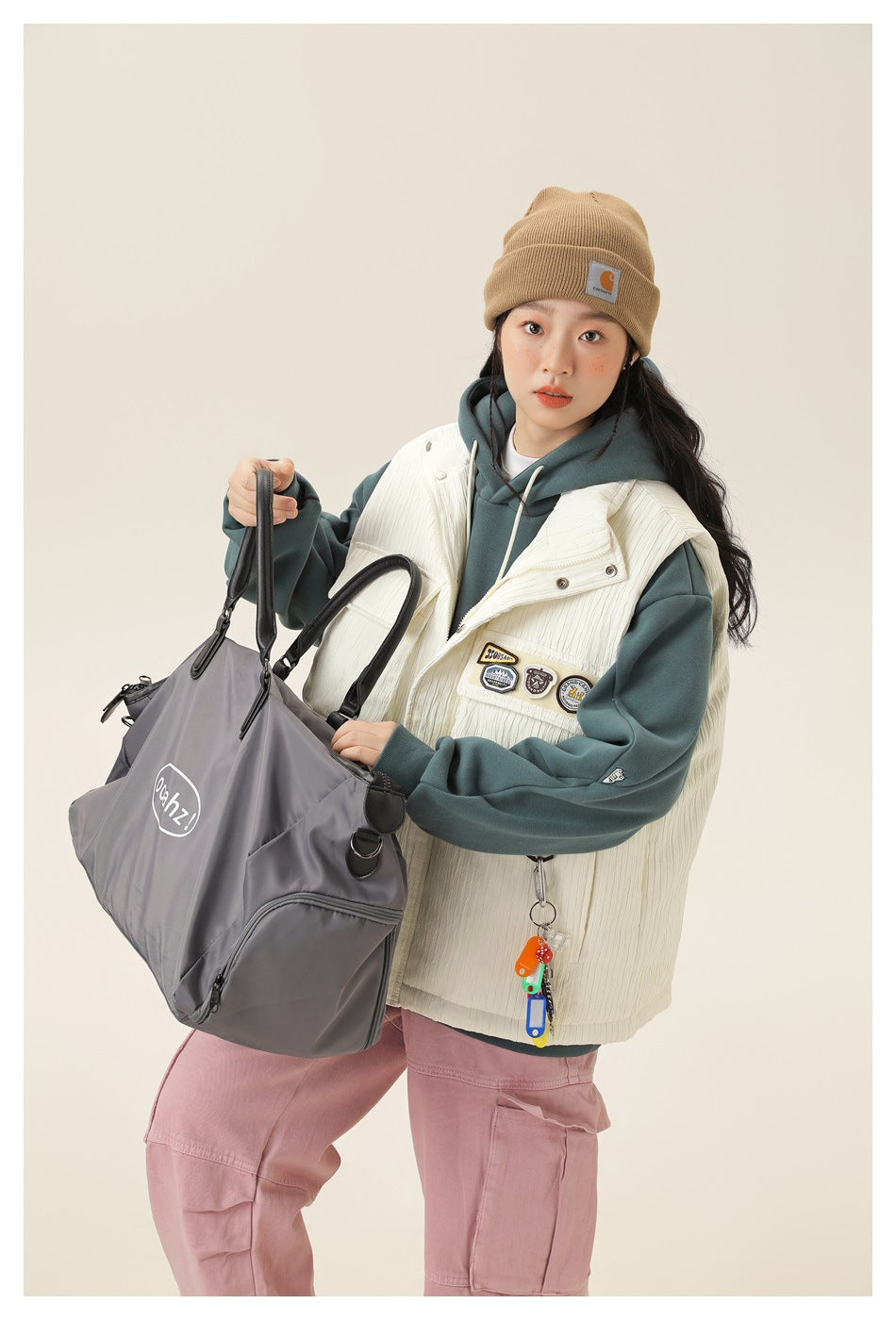 Model wearing a cream utility vest over a teal hoodie with pink cargo pants, holding a gray duffel bag.