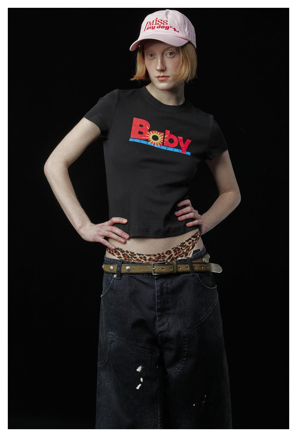 Model wearing a black graphic T-shirt with wide black jeans and a pale pink cap, hands on hips.