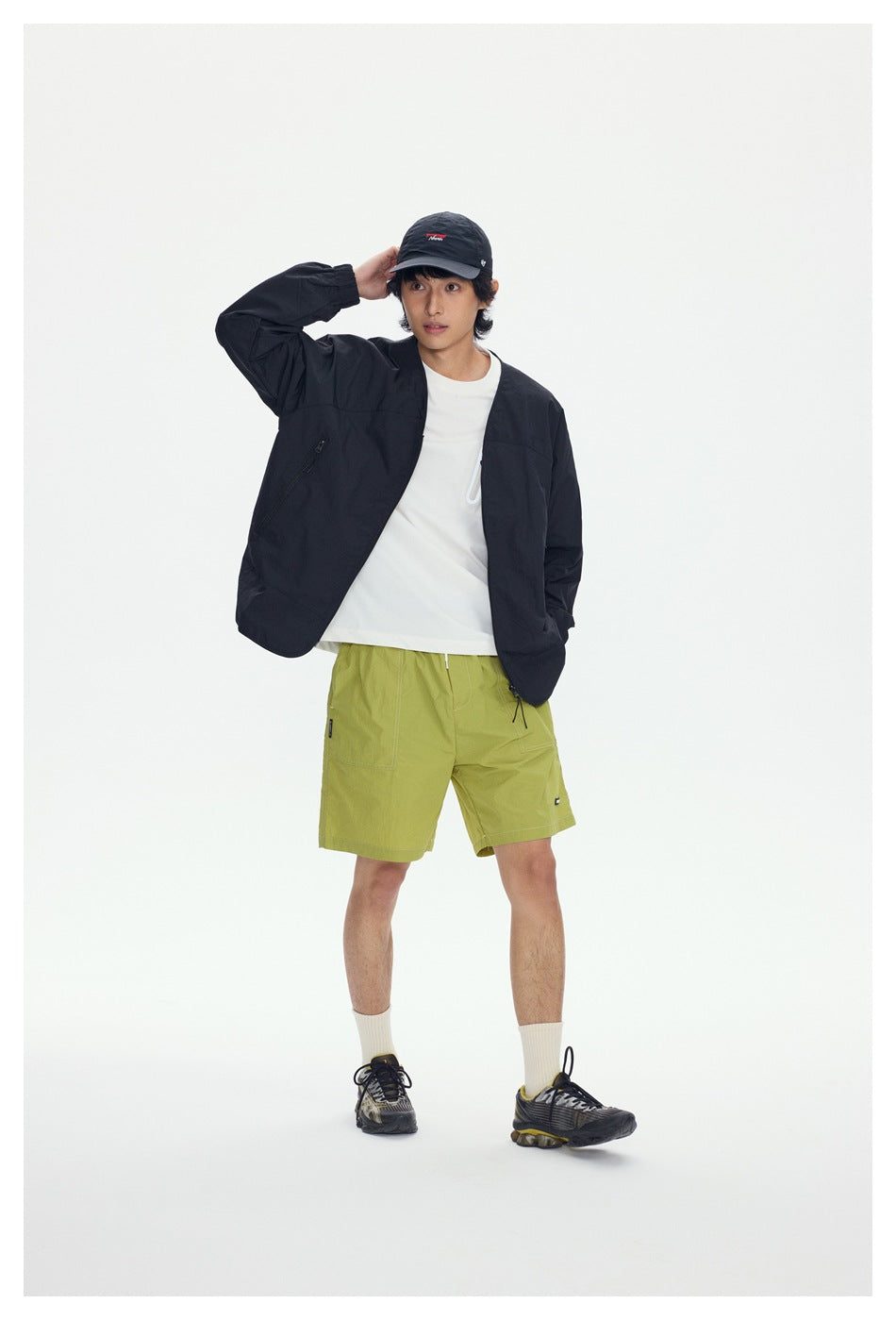 Model wearing an open black V-neck jacket over a white tee with green shorts, cap and sneakers, standing front view