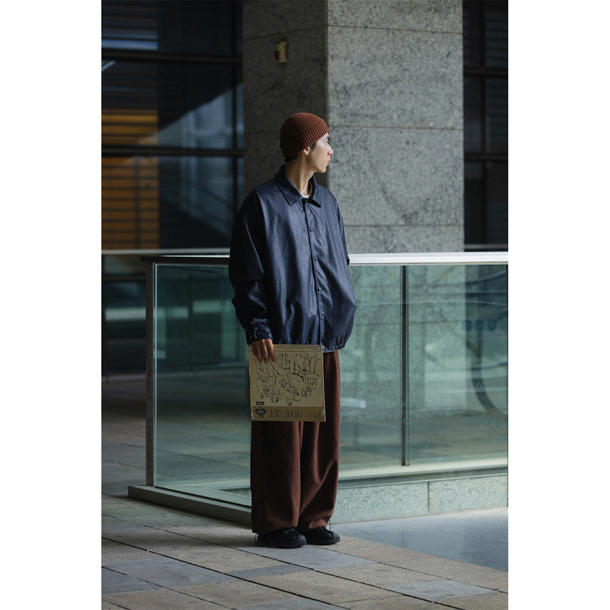 Model wearing an oversized navy button-front jacket and brown trousers, viewed full length outdoors.