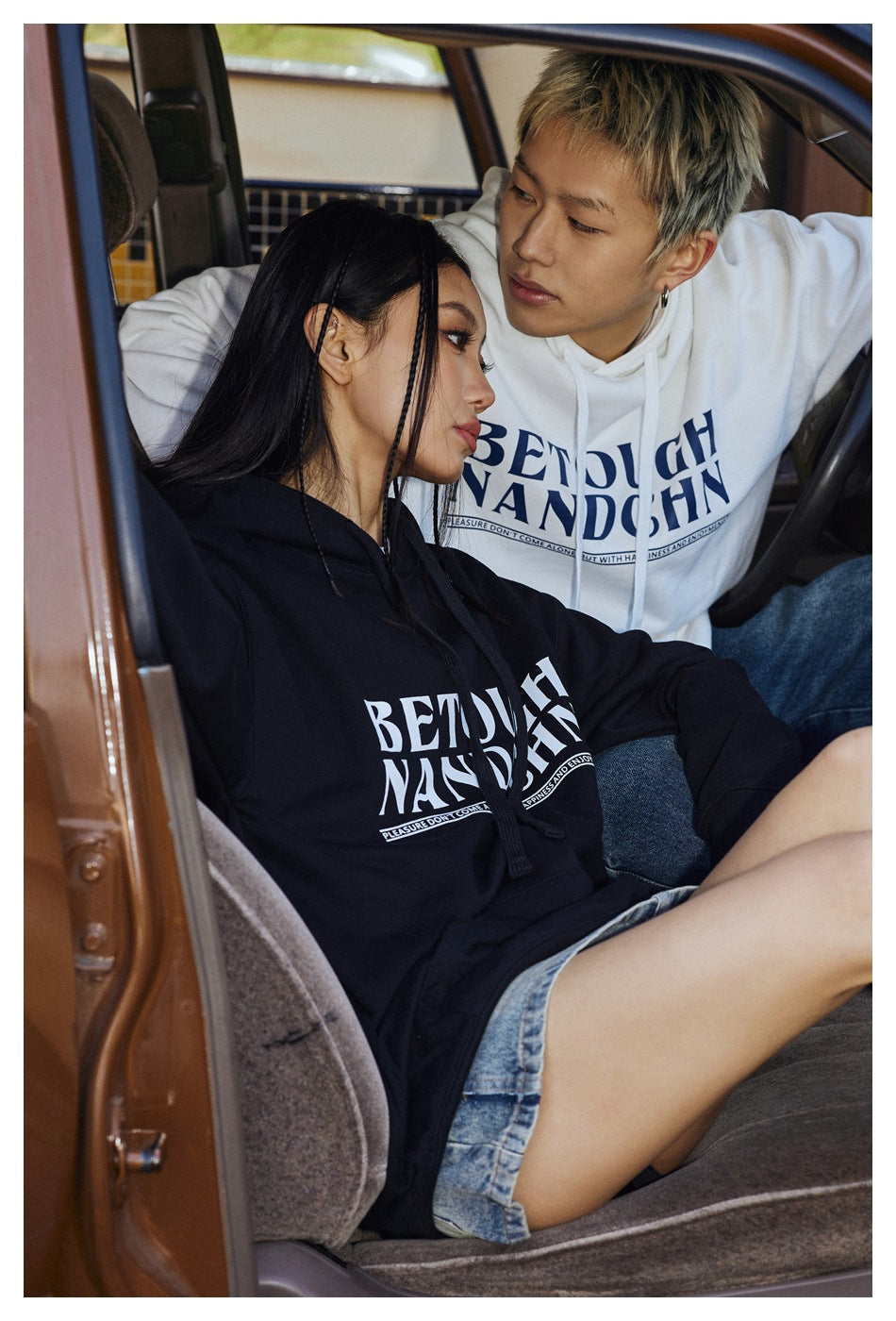 Seated models wear black and white oversized graphic hoodies, shown close up inside a car.