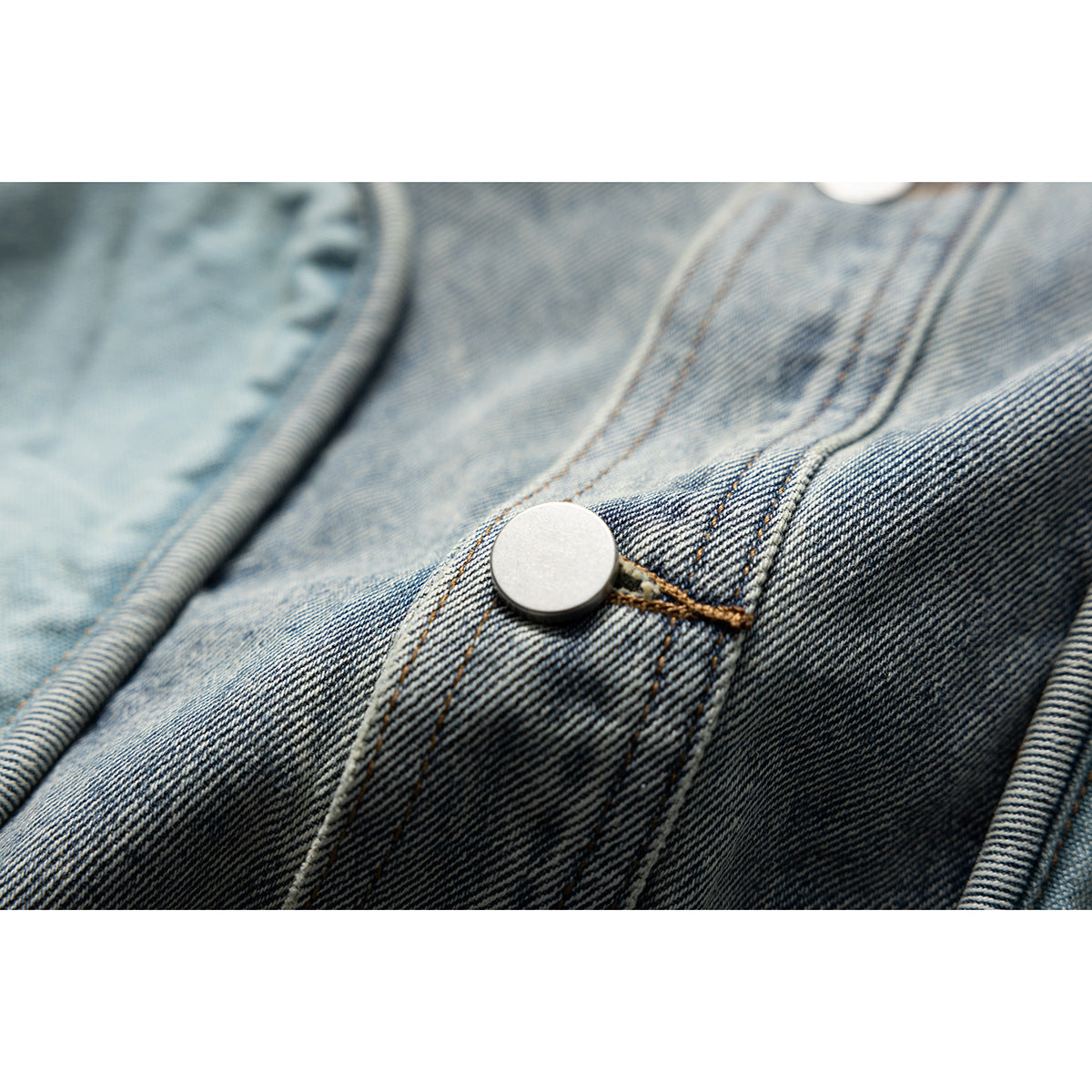 Macro view of a silver metal button and brown stitching on blue denim jacket fabric.