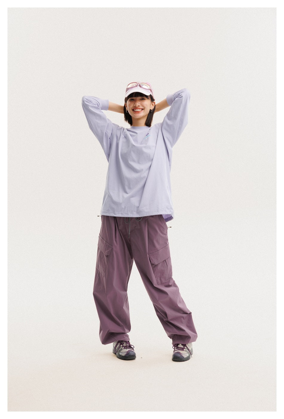 Model wearing a loose lavender long-sleeve shirt and mauve cargo pants, posing with hands behind her head.